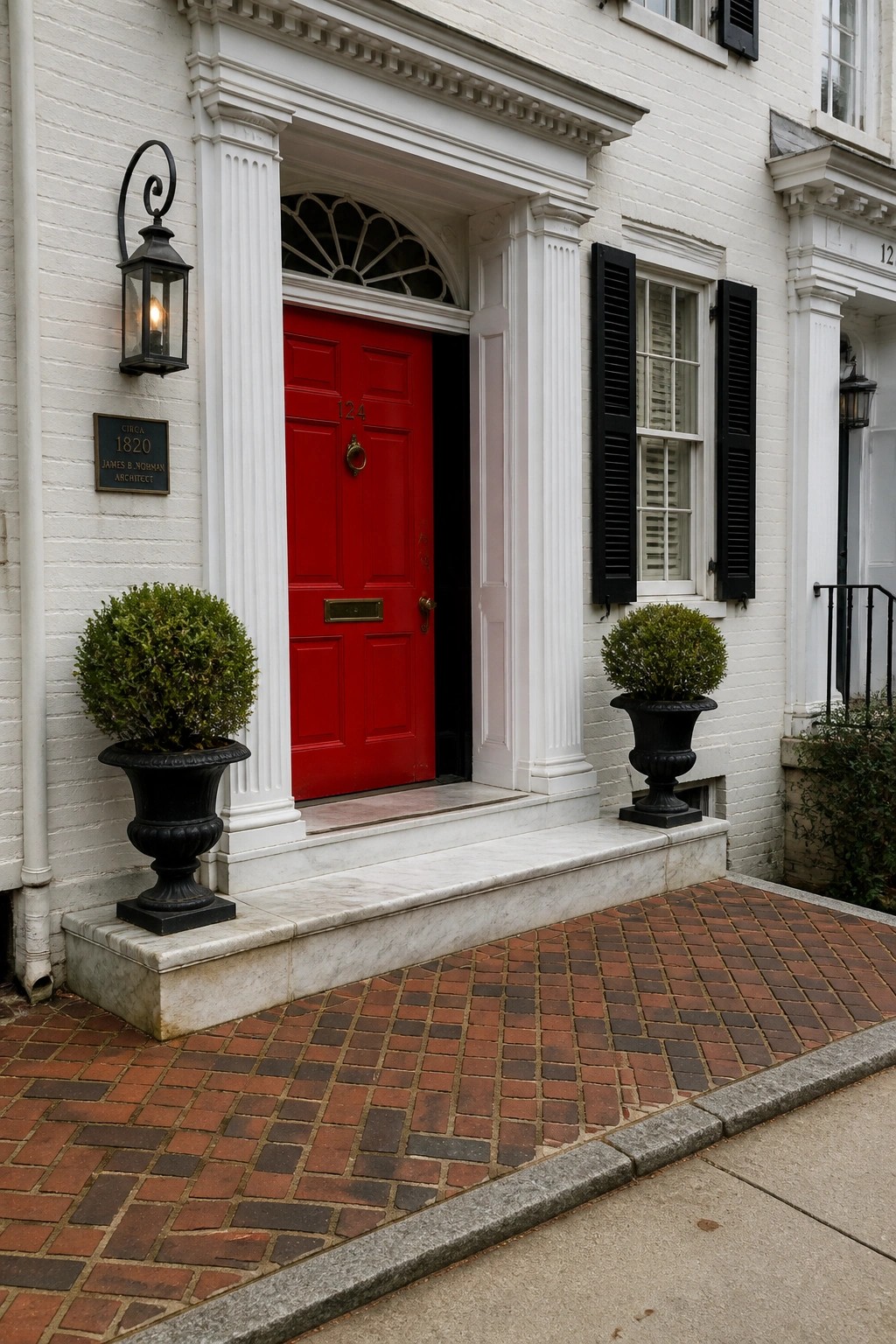 A bright red door centered in a white brick entry with white columns, a fanlight window above, and two black urns with shrubs on the steps.