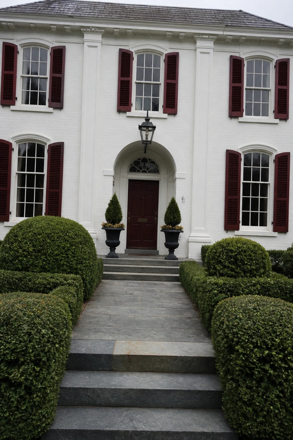 A white brick house exterior with deep red shutters, a centered red door under an arched opening, and two potted shrubs flanking the steps.