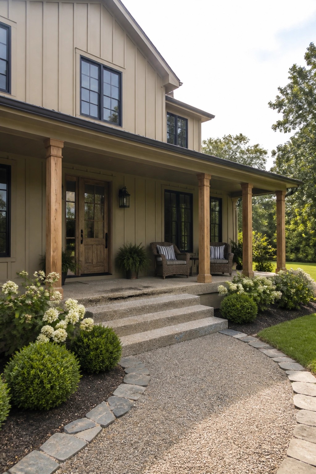 A beige house exterior featuring black-framed windows, a wooden porch with columns, concrete steps, and a gravel driveway lined with shrubs.