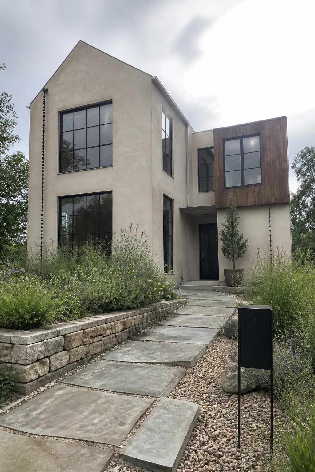 A modern two-story house with beige stucco walls, black-framed windows of varying sizes, and a stone retaining wall beside a stone paver walkway.