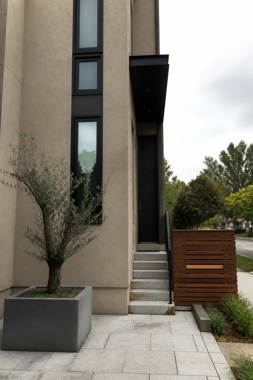 A beige stucco house exterior featuring tall black-framed windows, a black entry door, concrete stairs, and a large olive tree in a gray planter next to a horizontal wood screen.