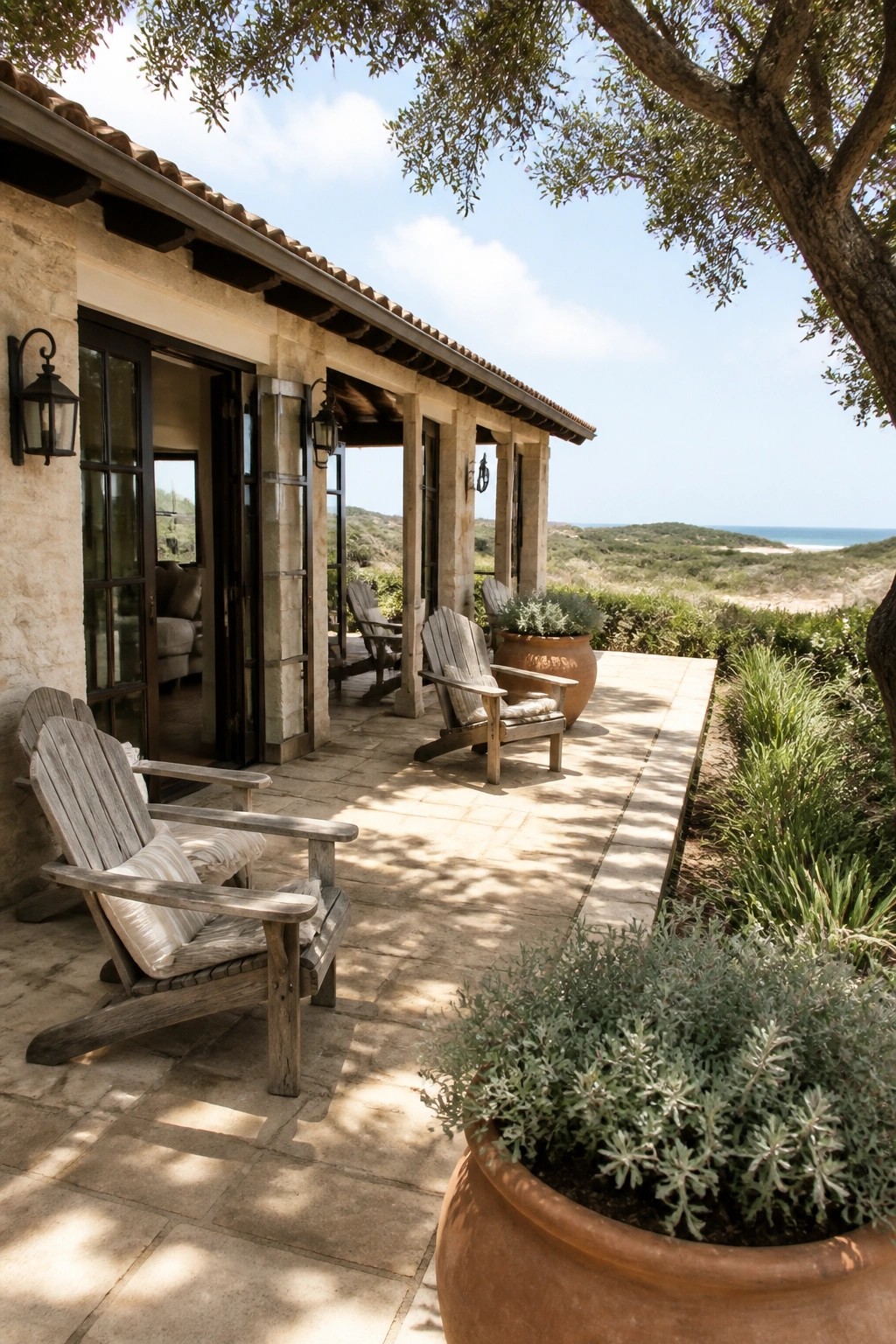 A stone house patio with several weathered wooden Adirondack chairs arranged on large beige stone tiles, next to terracotta pots and overlooking coastal hills.