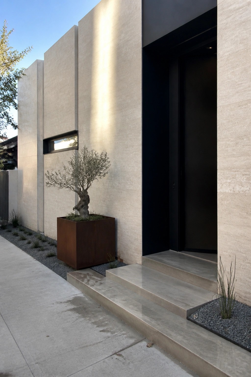 A beige stone house exterior with a black door, concrete steps, and a rusted metal planter holding a small tree beside the entry.