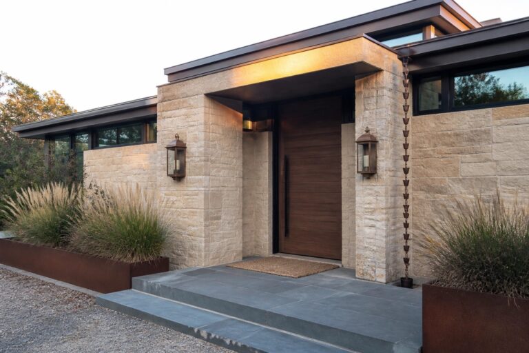A beige stone house exterior with a tall wooden front door, dark metal overhang, wall lanterns, and rusted metal planters holding tall grasses.
