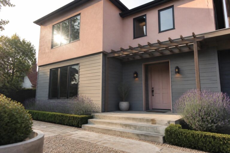 A two-story contemporary house with pink stucco on the upper level, gray siding below, black-framed windows, a pink front door, and concrete steps leading to the entry.