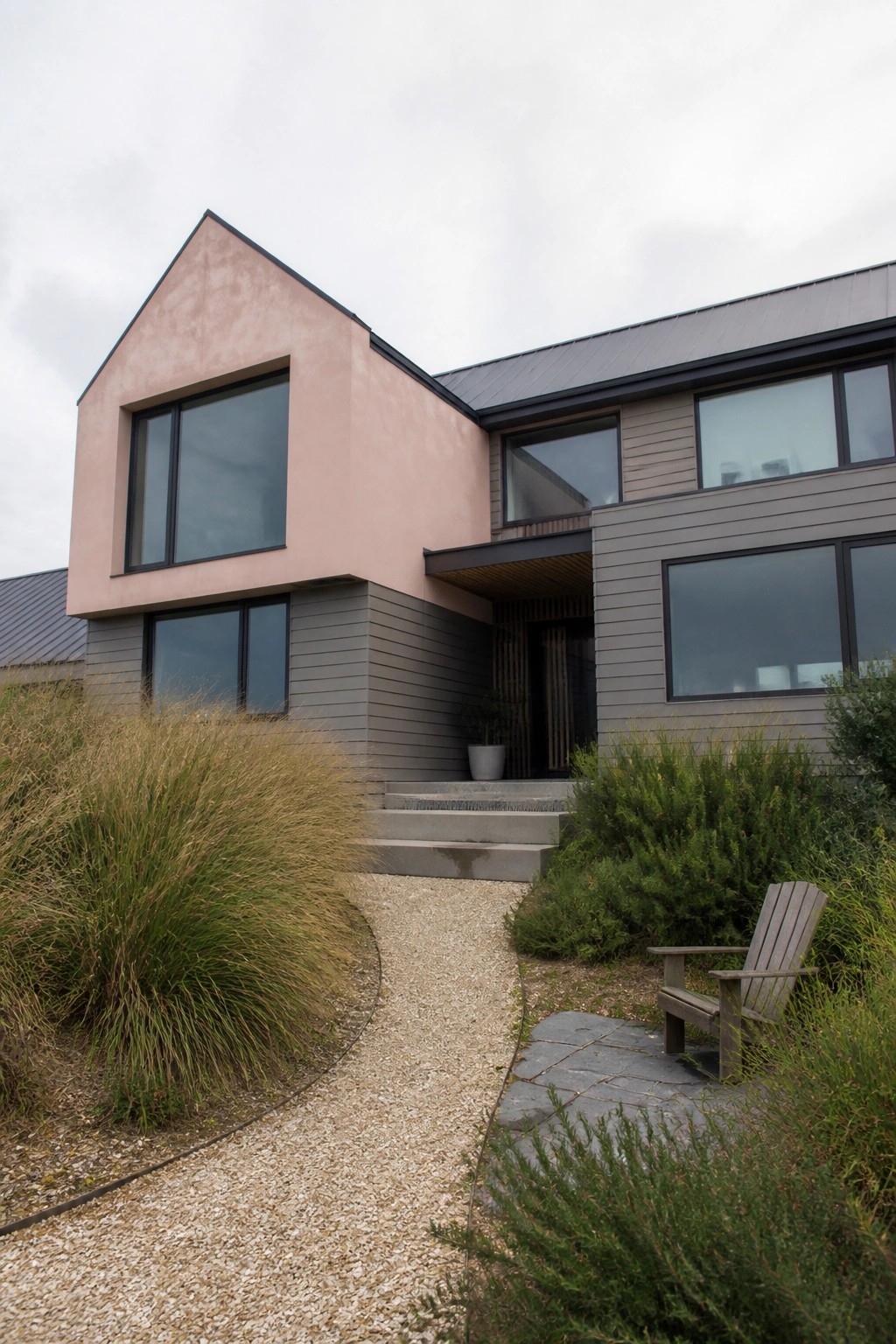 Modern pink-and-gray house with large windows, gravel path, wooden chair, and lush landscaping.