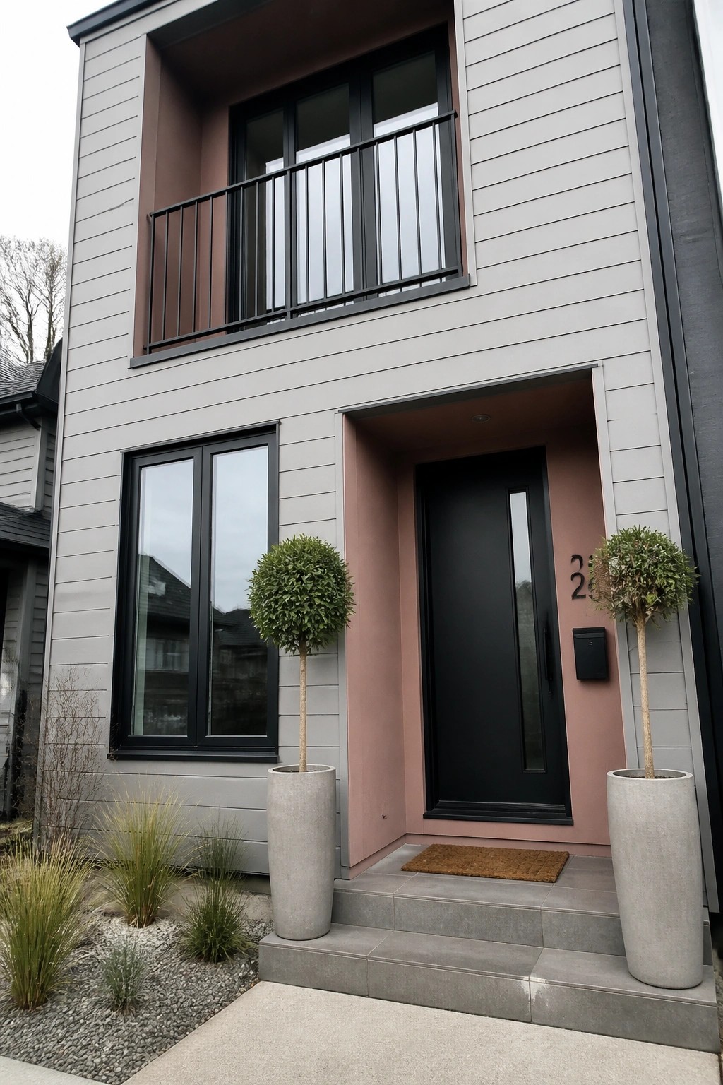 Modern gray house with black door in pink alcove, flanked by potted topiary plants.