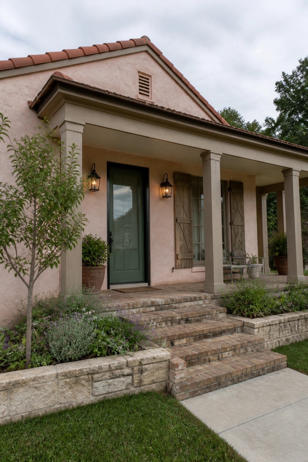 A soft pink stucco house with red clay tile roof, green front door, wooden shutters on the windows, brick steps, and a covered porch supported by columns.