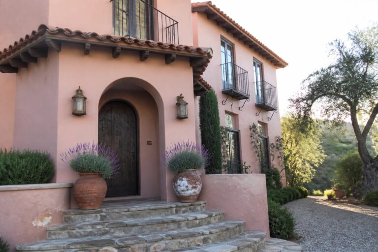 Pink stucco house with arched wooden door, stone steps, and potted lavender plants.