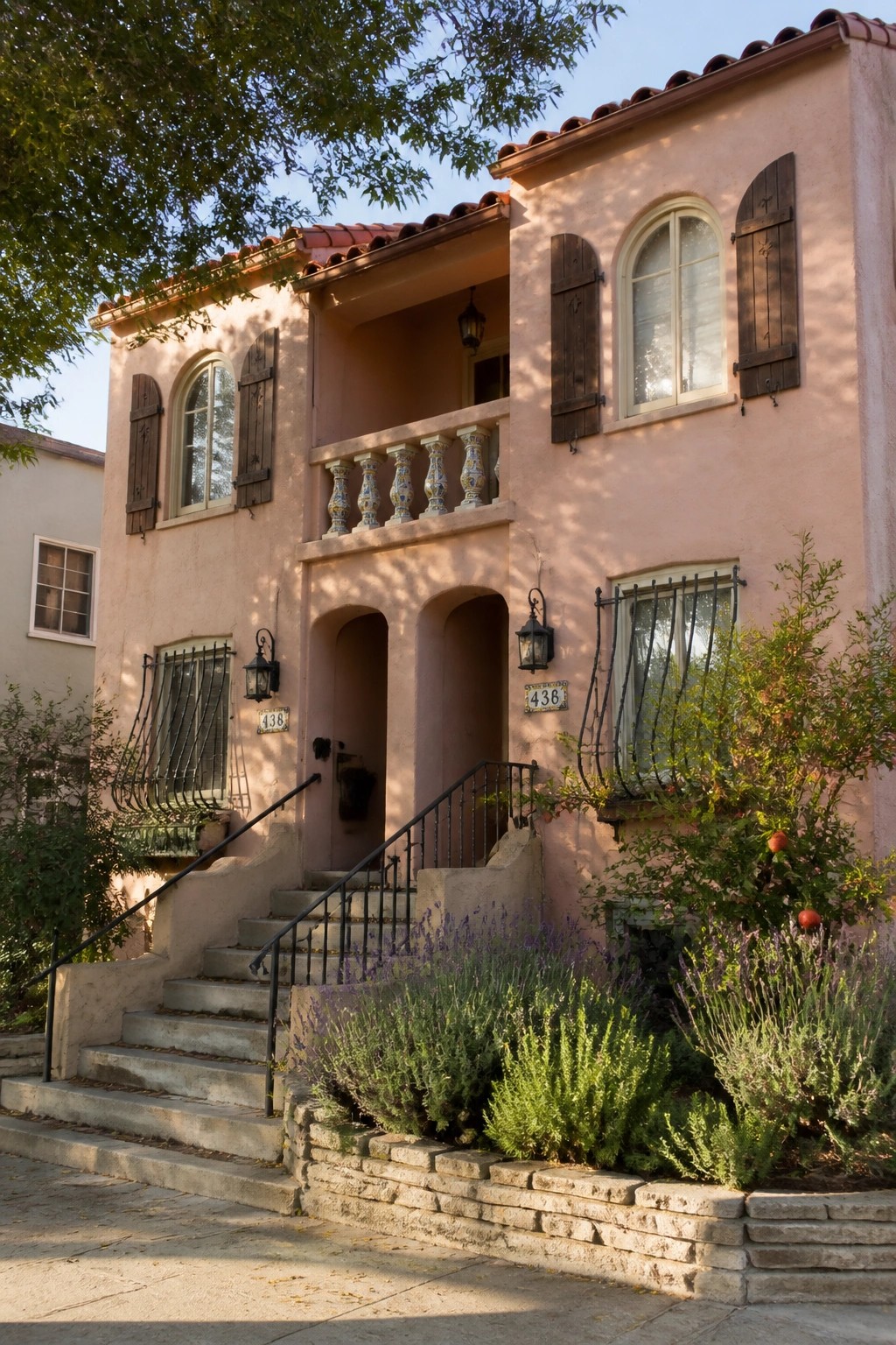 A two-story pink stucco house with a red clay tile roof, arched windows with dark shutters, a small balcony with colorful balusters, and concrete stairs leading to the front entry.