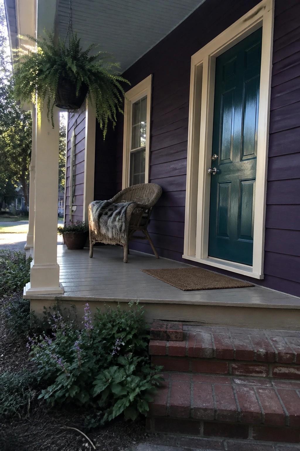 Purple house porch with teal door, wicker chair with blanket, hanging fern, and brick steps