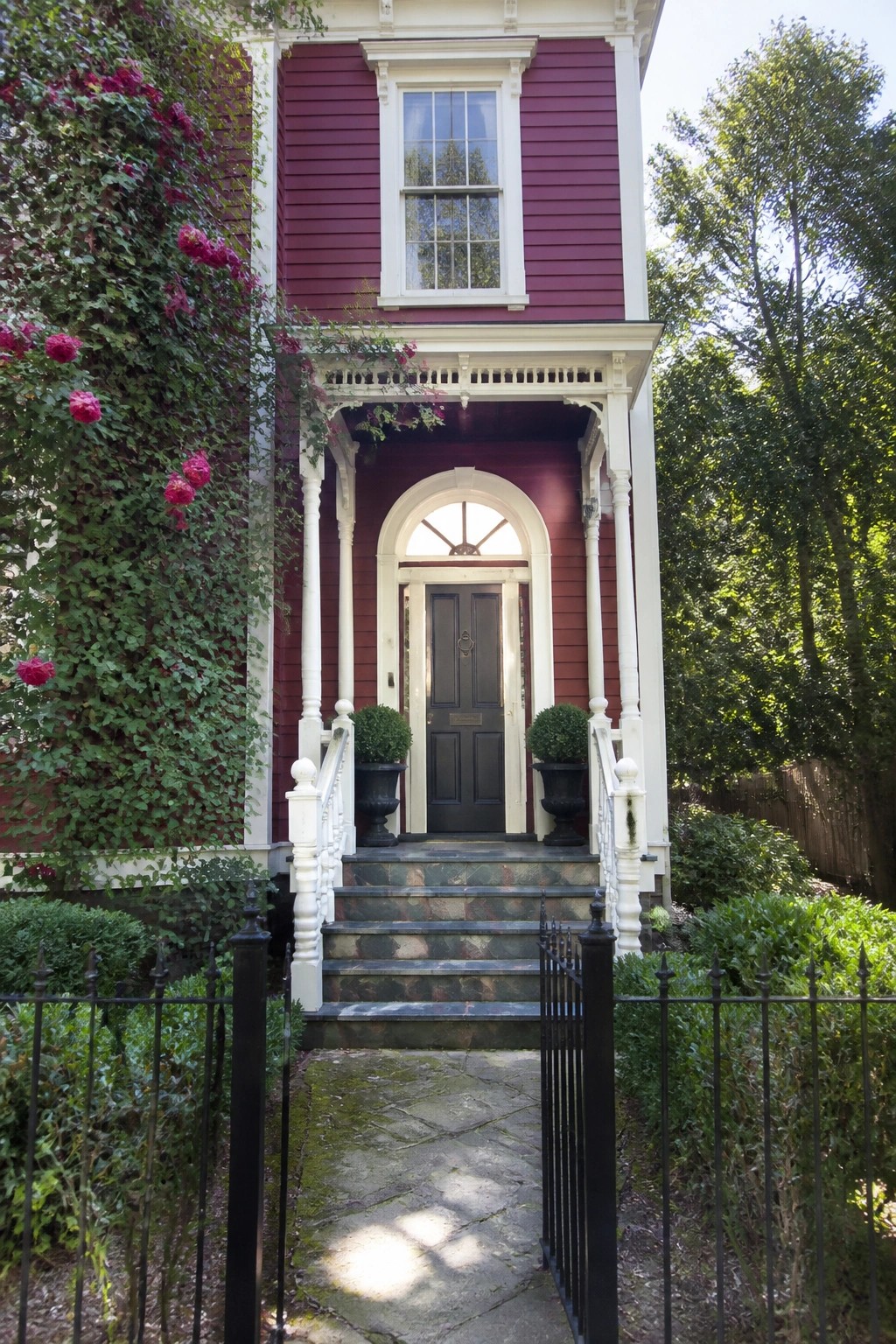 A two-story house with dark red siding, white trim, columns, and steps leading to a dark front door, set among trees and shrubs.