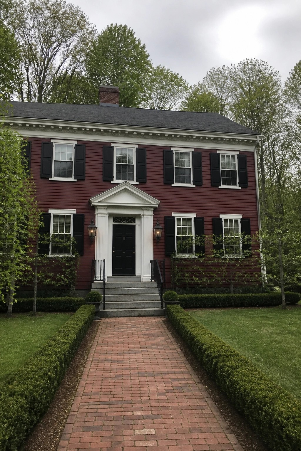 A two-story dark red house with white trim, black shutters, a central white portico, and a brick walkway leading to the front steps, set among trees and hedges.