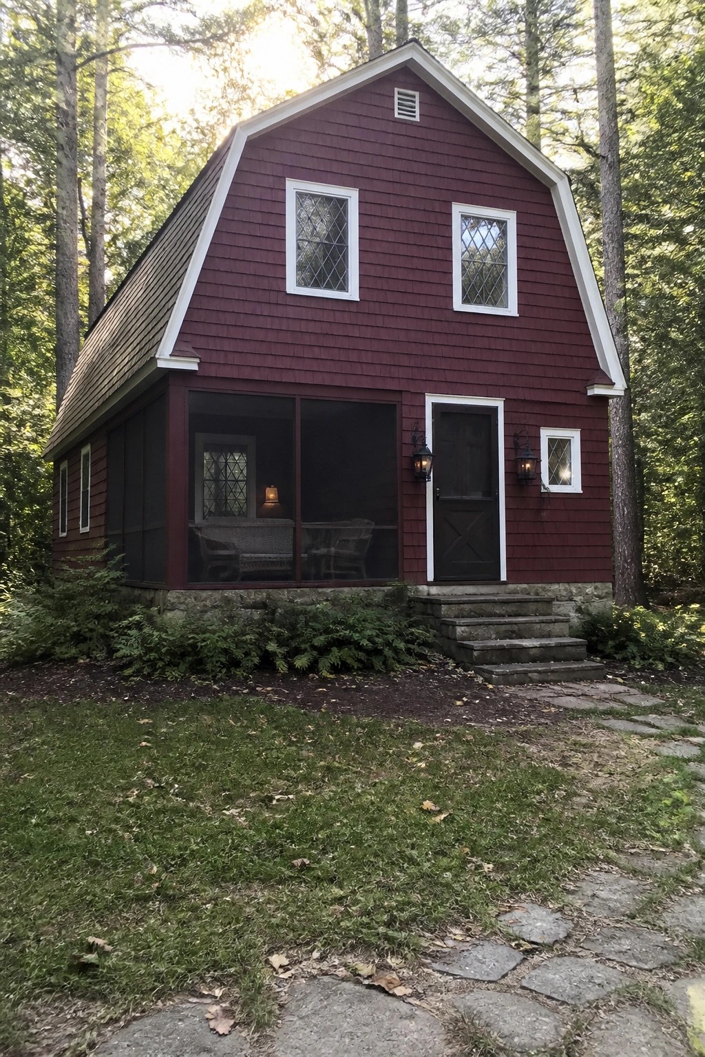 A dark red gambrel-style house with white trim sits among tall trees, featuring a screened porch, stone foundation, and steps leading to the front door.