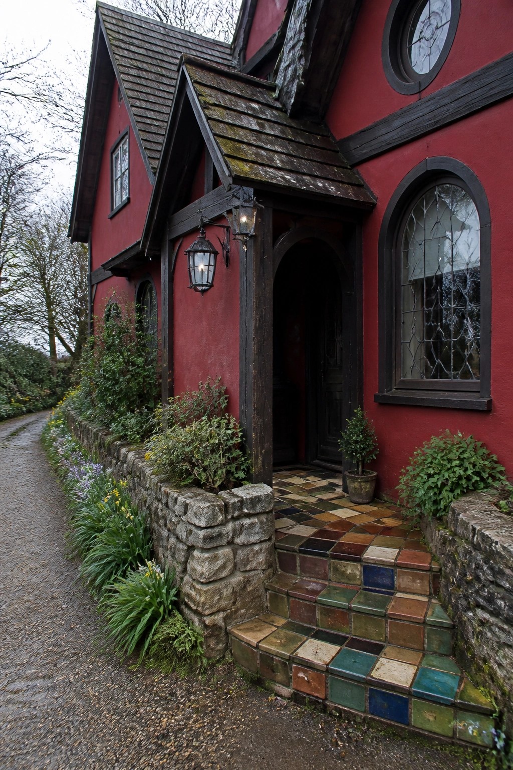 A red house exterior with dark wooden trim, an arched doorway, leaded glass windows, and colorful tiled steps beside a gravel path.