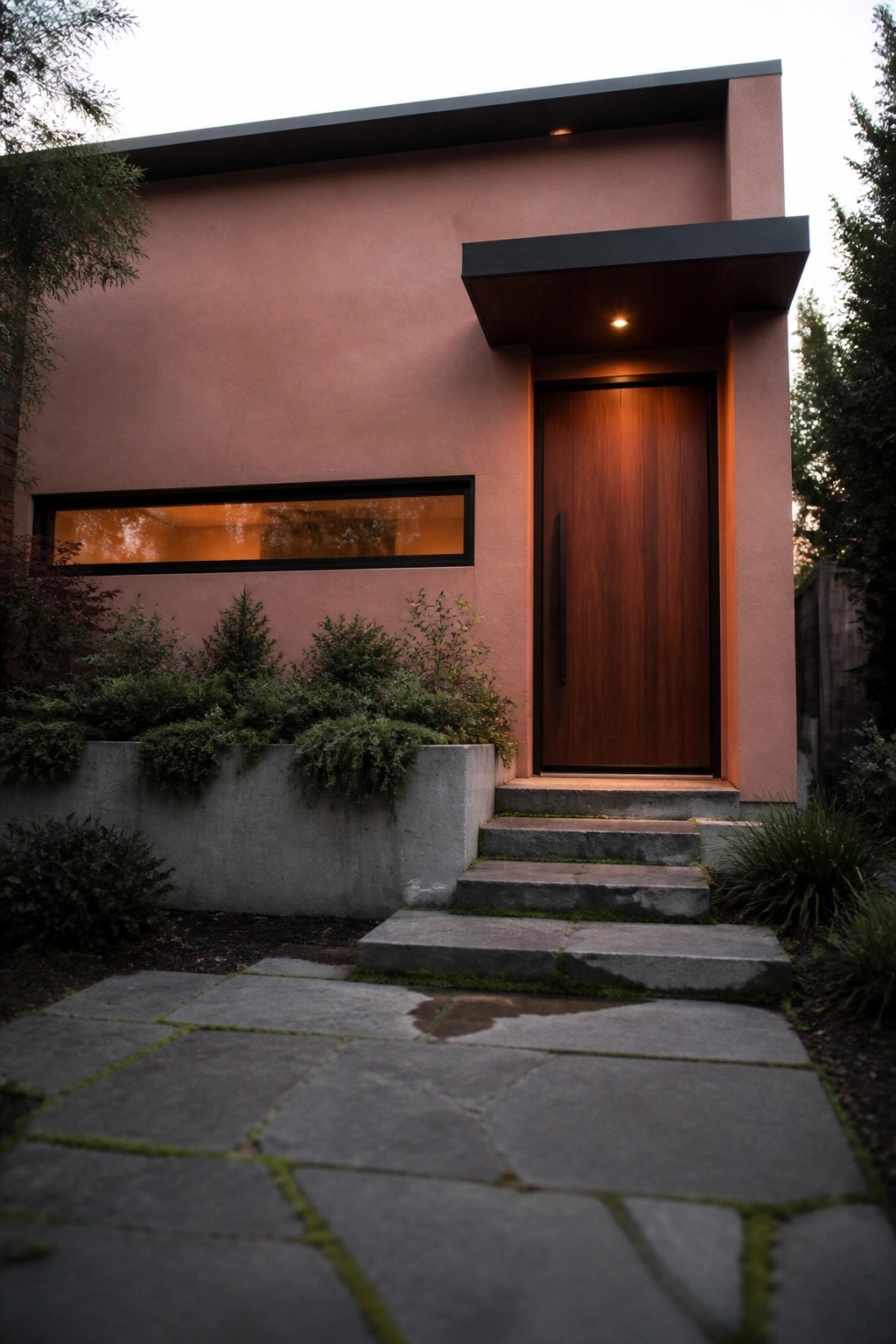 A modern dusty pink stucco house exterior with a tall vertical wood front door, dark overhang, concrete steps, and a narrow horizontal window.