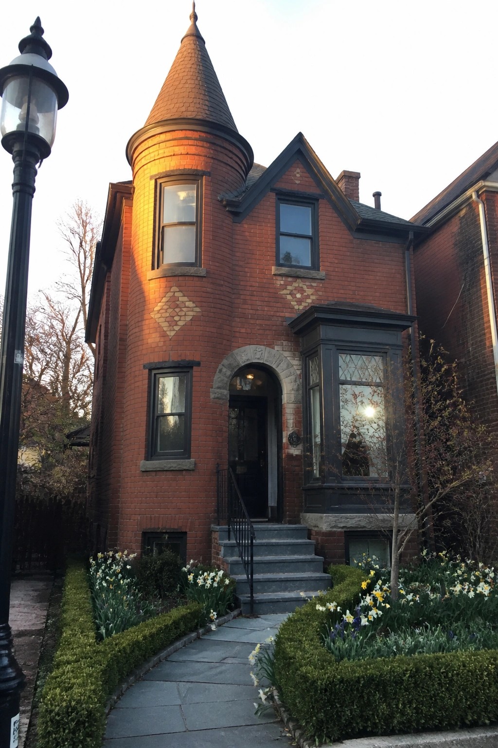 A red brick Victorian house featuring a round turret, arched stone entry, and a stone path lined with flowers leading to the front steps.