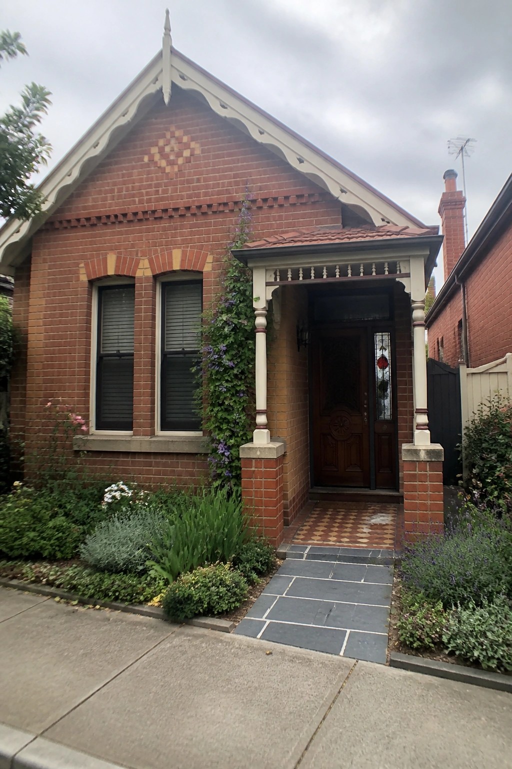 A red brick Victorian house facade with a decorative diamond brick pattern in the gable, two front windows, a wooden door under a porch with white columns, and a tiled entry path.