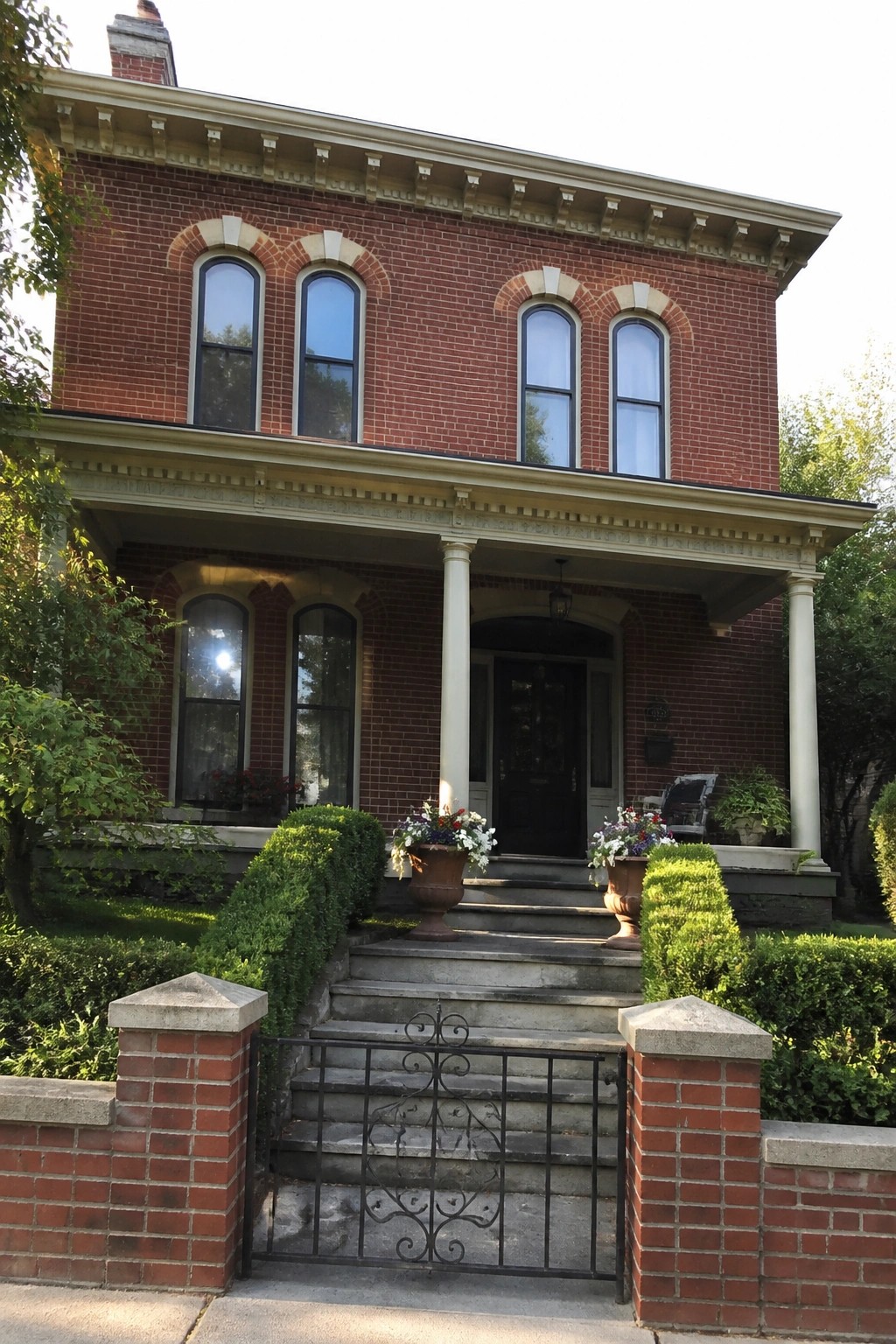 A two-story red brick Victorian house with cream painted trim around the windows, cornice, and porch columns, a dark front door, and stone steps with a black metal gate.