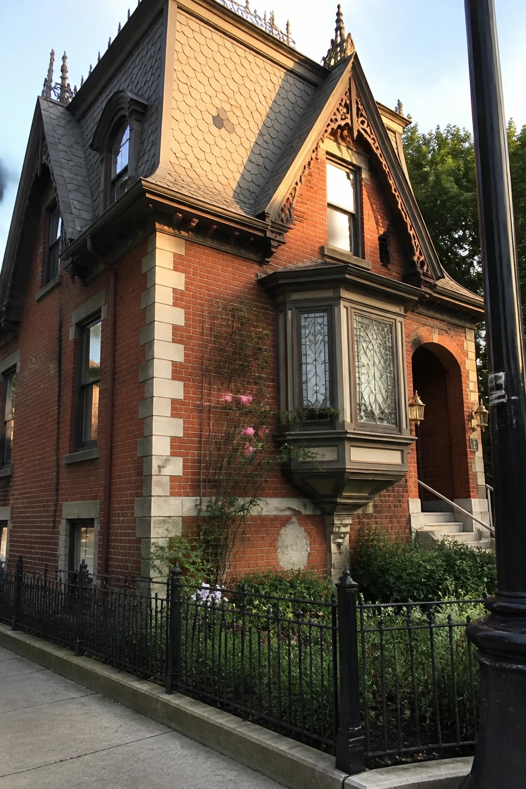 Ornate Victorian red brick house with steep slate roof, bay window, and iron fence.