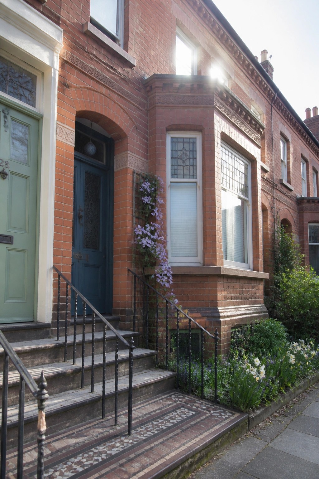 A red brick Victorian row house with a dark blue door under an arch, white-framed windows with decorative brick and stone detailing, black metal railings on the steps, and purple flowers growing up the side of the entrance.