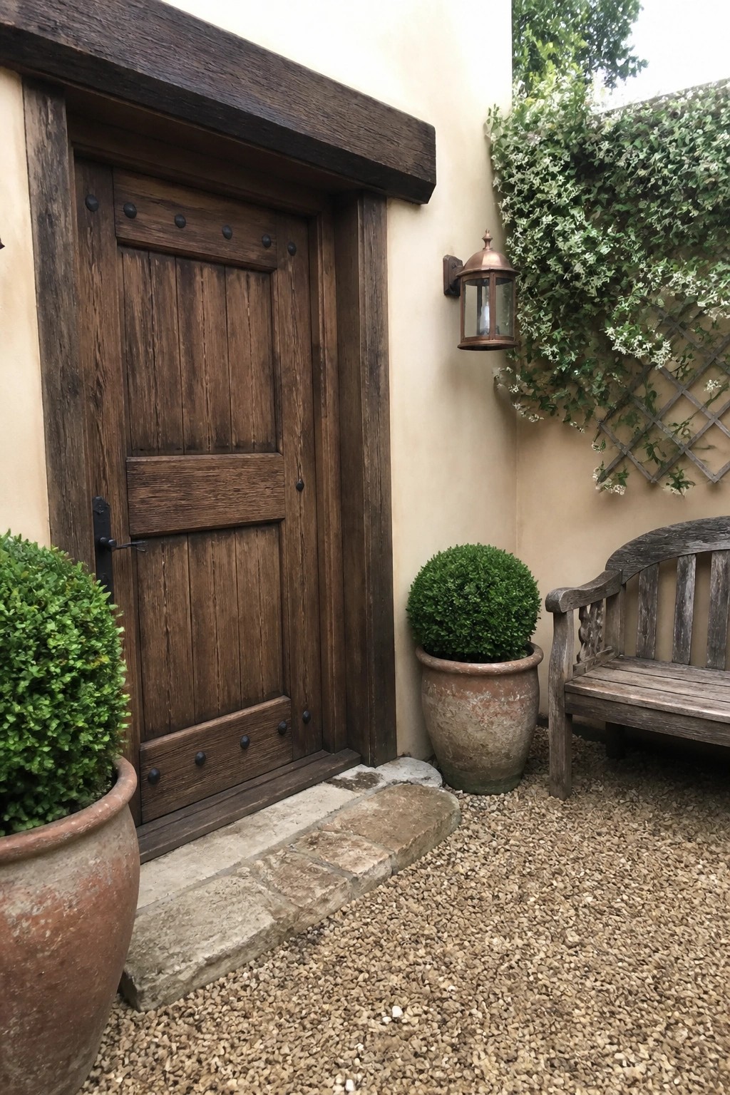 A dark wooden door in a beige stucco wall next to gravel ground, terracotta pots, and a wooden bench.