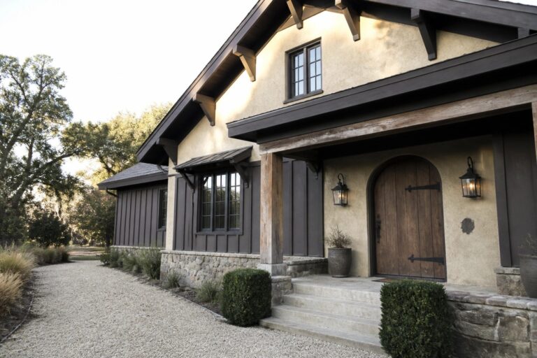 A two-story house with beige stucco walls, dark wood trim and beams, an arched wooden front door, and a stone base along the entry steps.