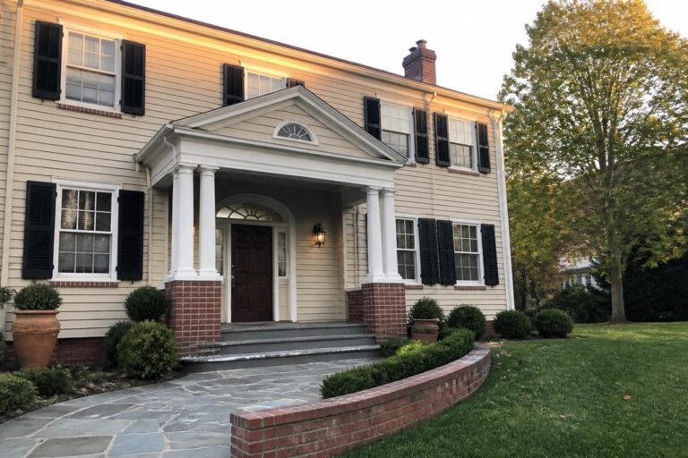 A two-story beige house with red brick foundation, white entry columns, dark shutters, brick steps, and a curved brick-edged path.