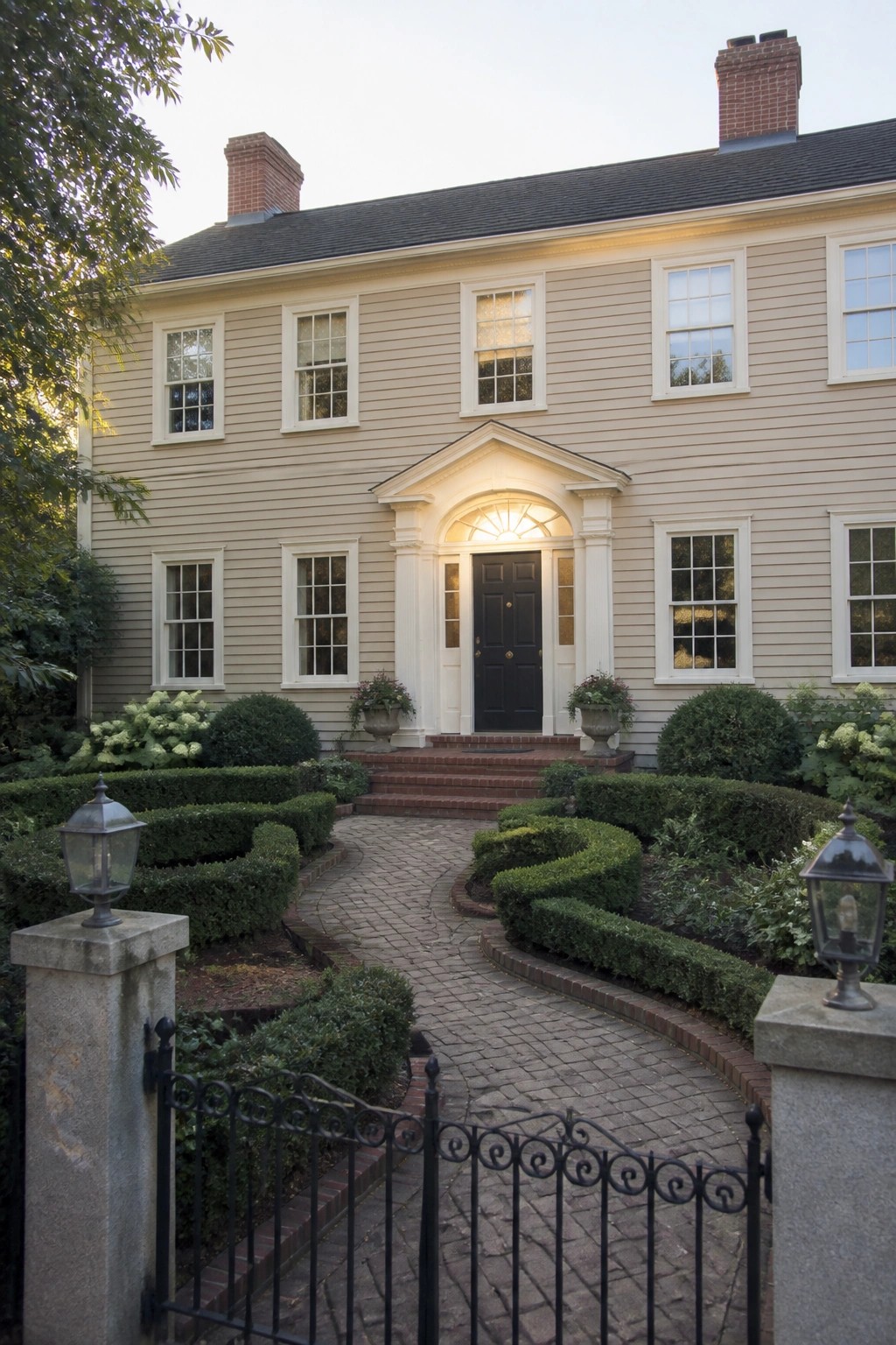 Two-story beige colonial house with black door, brick driveway, hedges, and iron gate