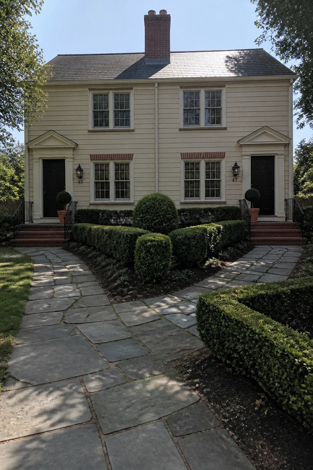 A two-story beige house with a red brick chimney, black front doors, white trim, and a curved stone path leading through the front garden.