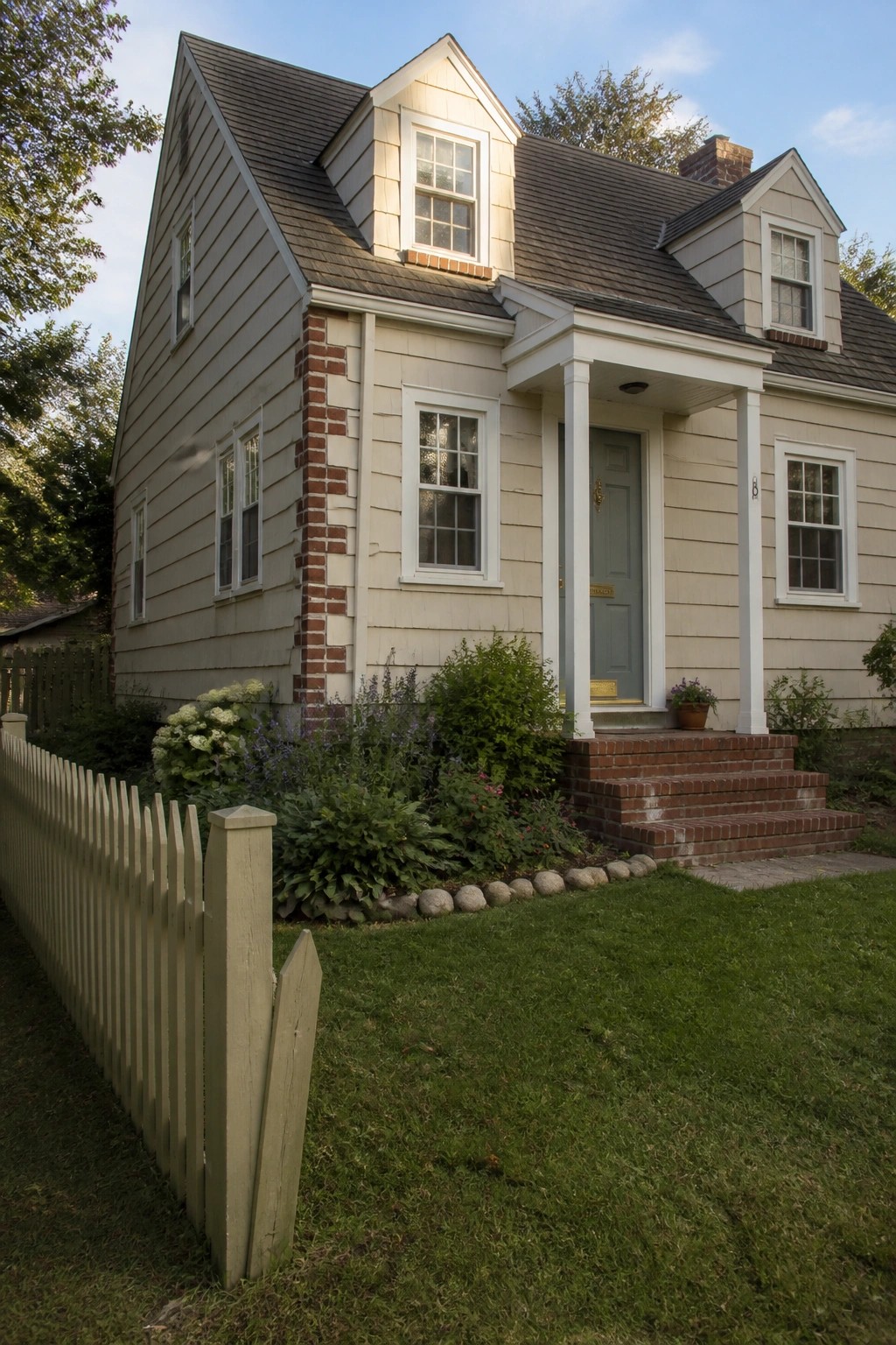 A two-story beige house with red brick accents around the corners and chimney, a white front porch with columns, gray door, and brick steps.