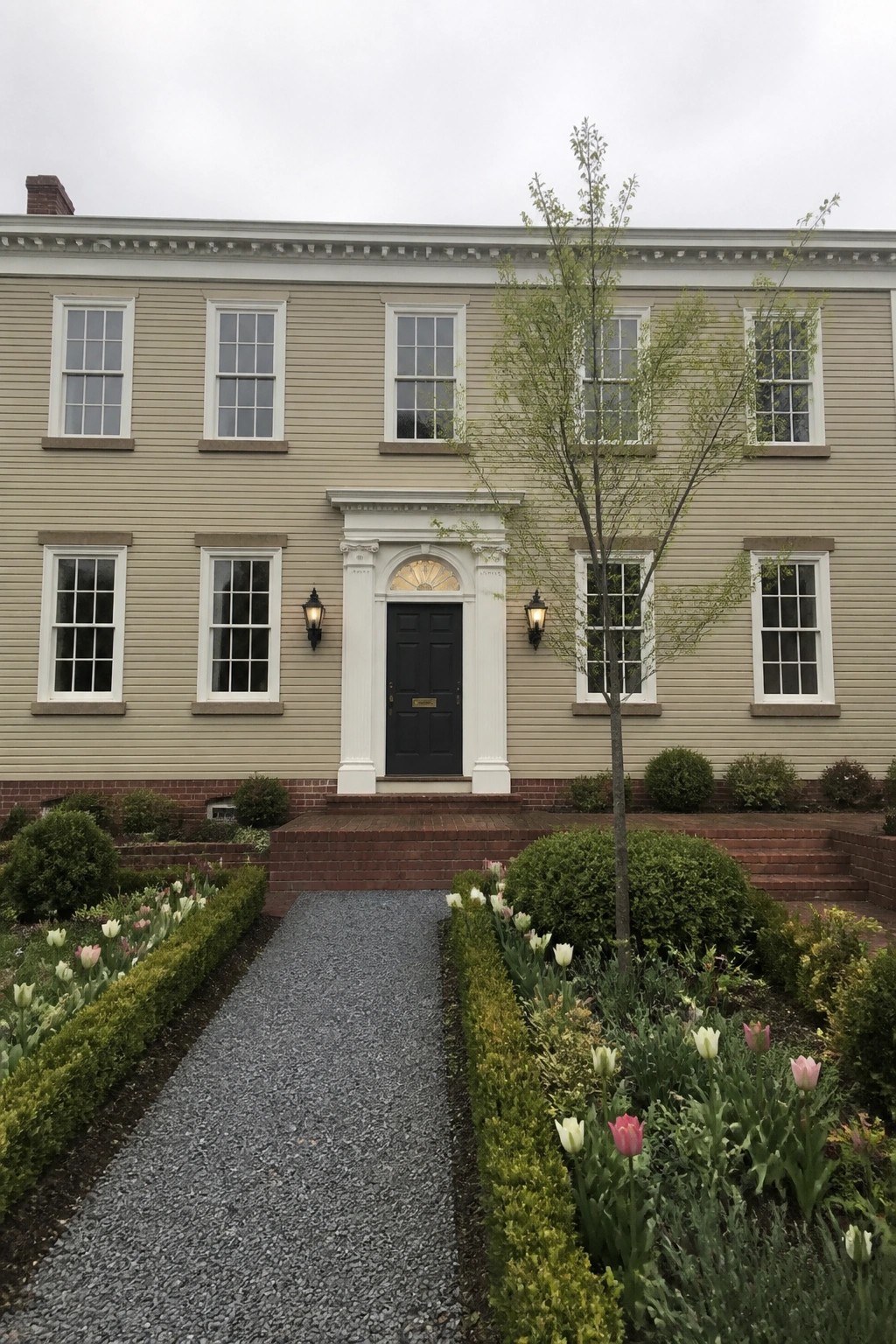A two-story beige house with white trim, a central black door under a white portico, red brick foundation and steps, and a gravel path lined with tulips and shrubs.