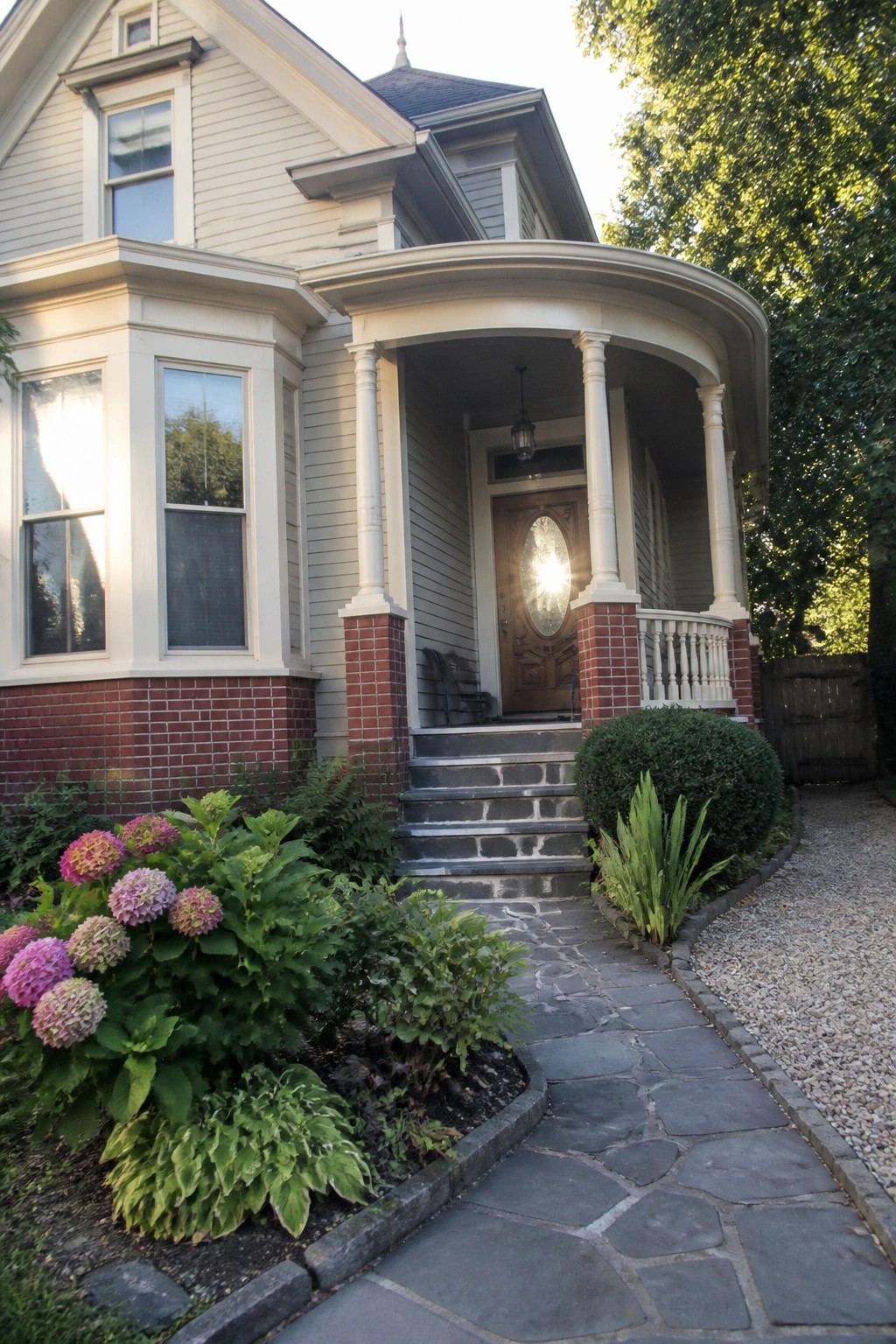 A beige traditional house exterior with red brick on the lower walls and porch piers, a curved white porch, stone steps, and a wooden front door.