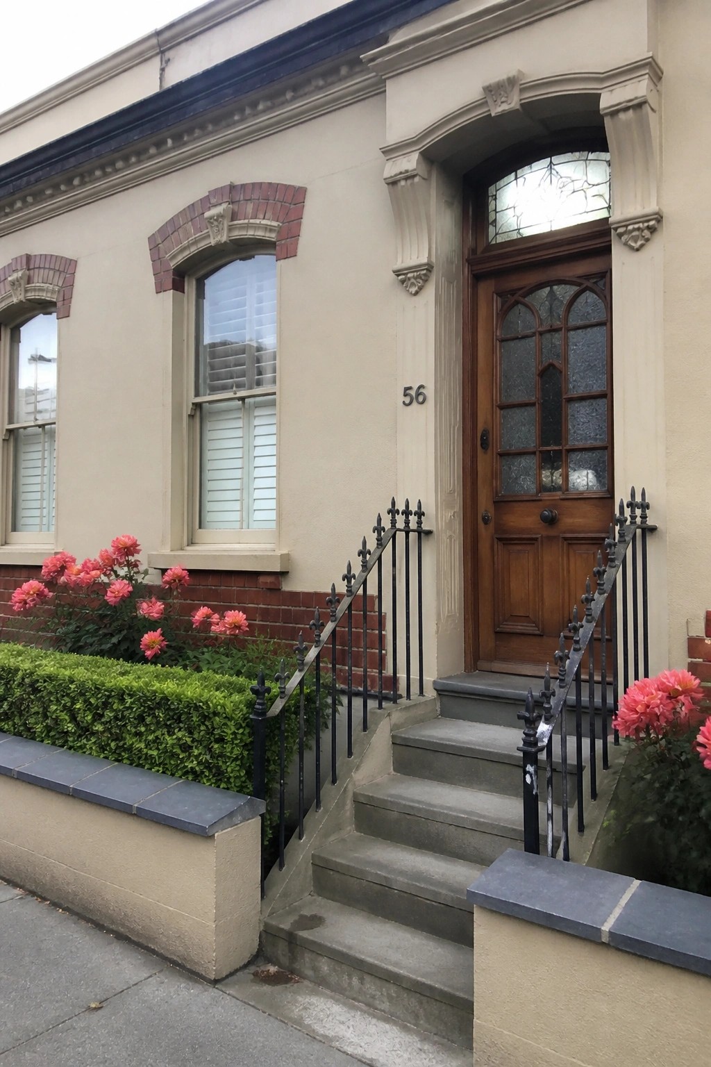 Beige house exterior with red brick accents around two windows, a wooden arched door, concrete steps, and pink flowers along the front.