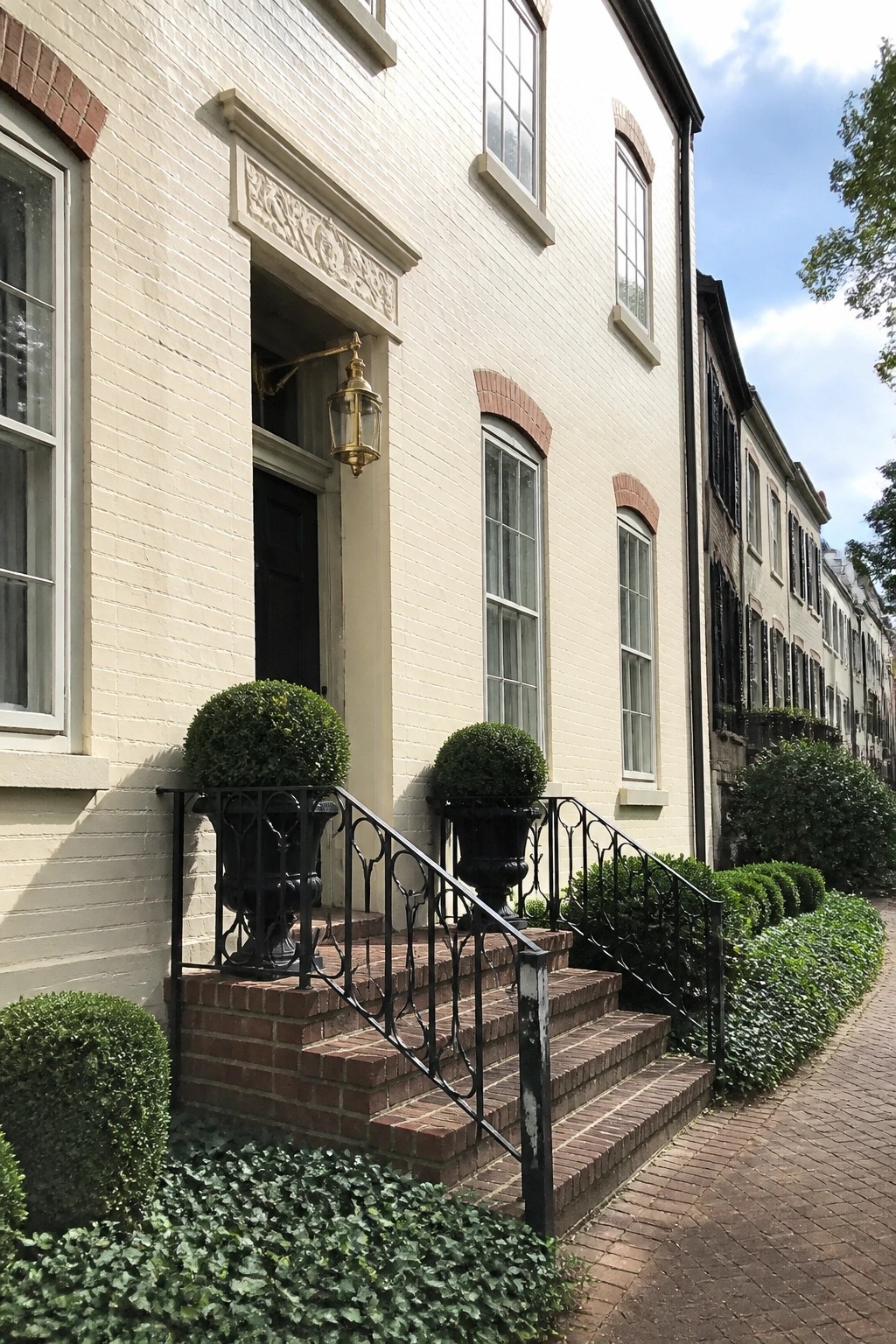 Beige brick house with red brick steps leading to a black door, black metal railings, and two large potted topiary plants on either side of the entrance.