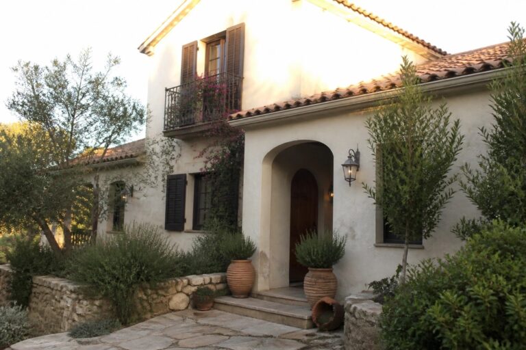 A cream stucco house with terracotta roof tiles, an arched front entry, a small balcony with railing, and stone steps flanked by potted plants.