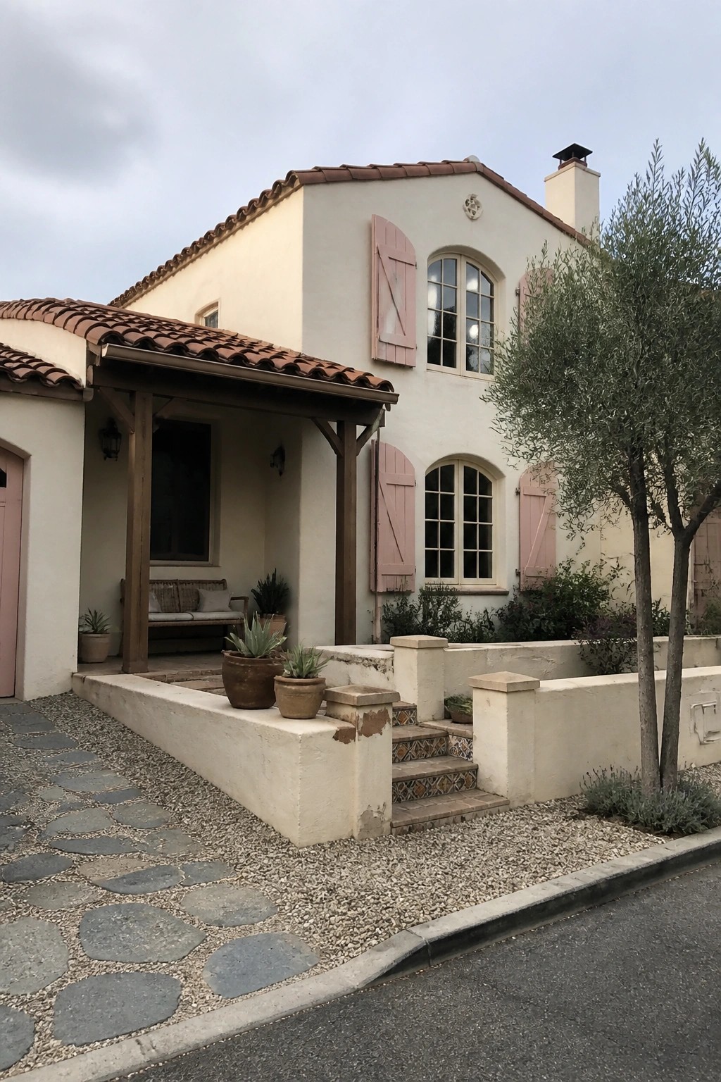 A cream stucco Mediterranean house with pink shutters, a terracotta tile roof, and a covered wooden porch.