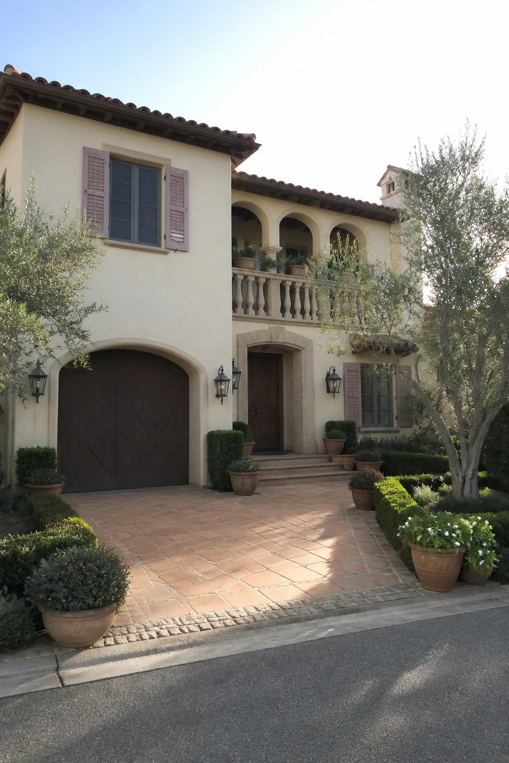 A two-story Mediterranean-style house with warm cream stucco walls, muted pink shutters, an arched wooden garage door, a stone balcony, and terracotta roof tiles.