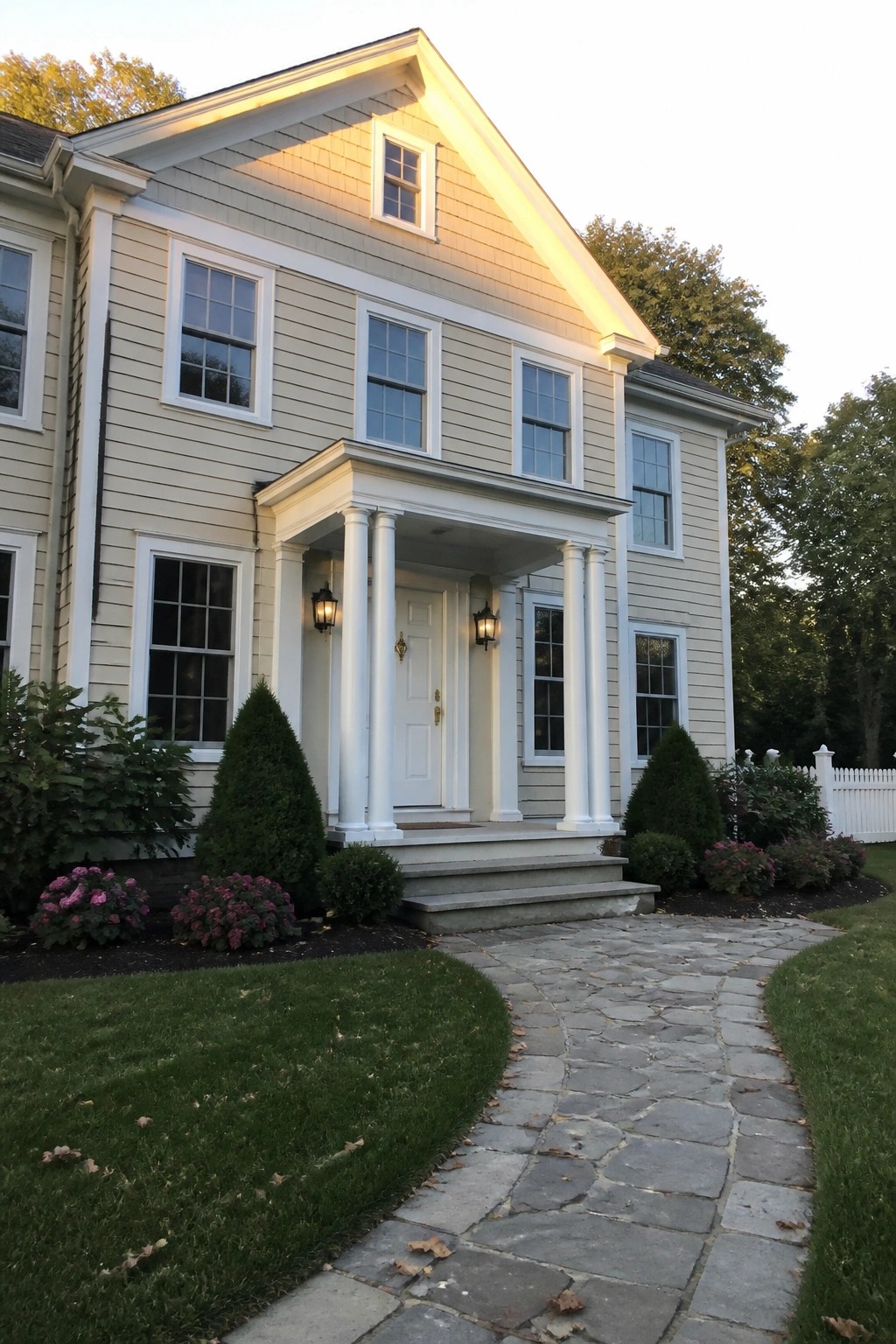 A two-story beige house with white trim, columns, and a stone path leading to the front entrance.