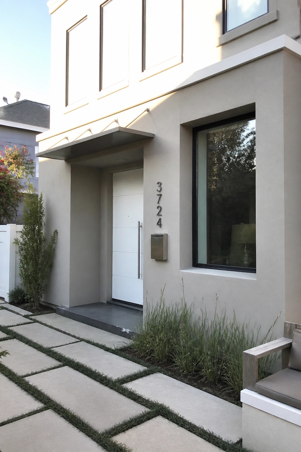 Front view of a modern two-story beige house with a white entry door, recessed porch, metal house numbers, and a concrete paver walkway with grass strips.