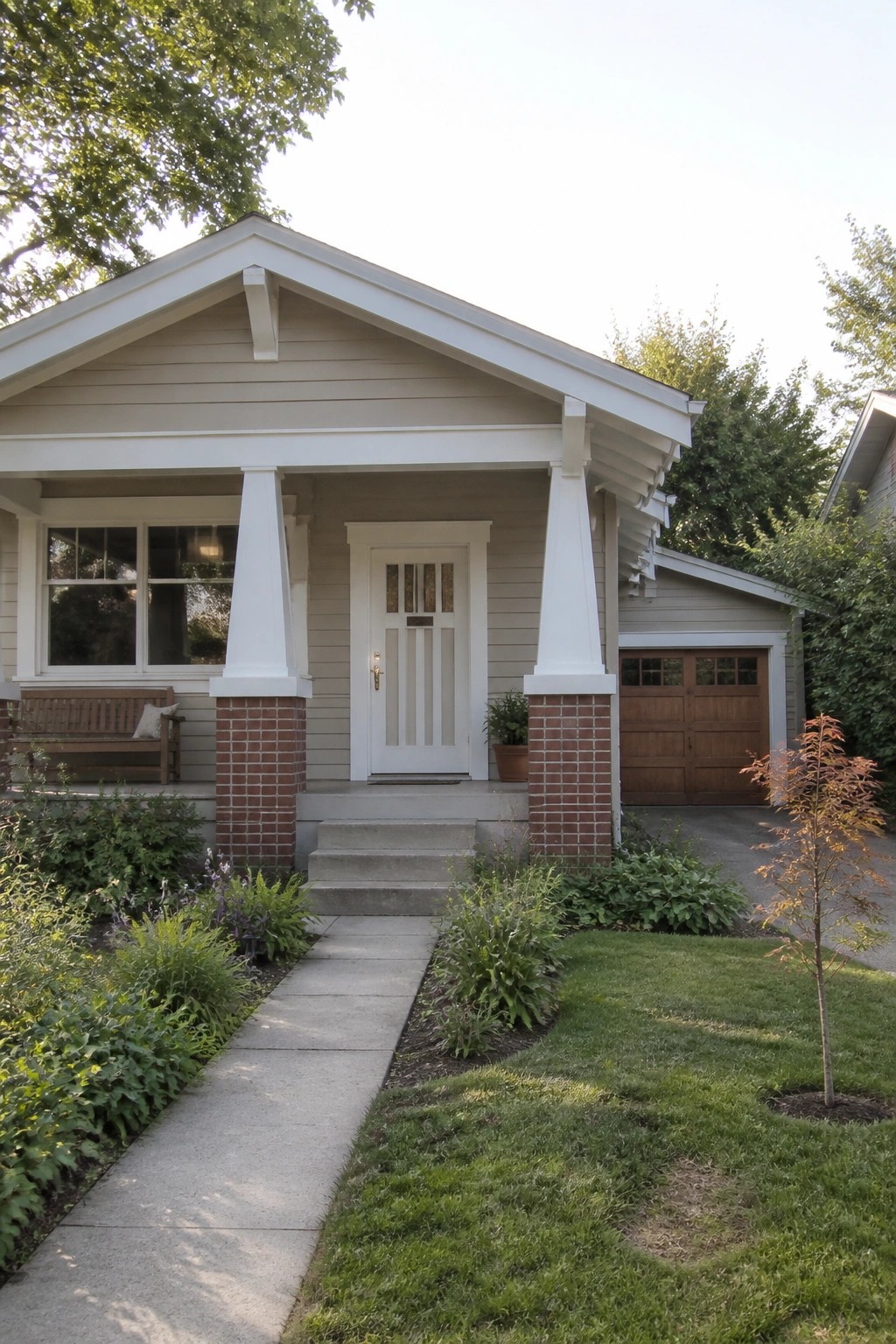 A beige house with white trim on the gable, porch columns, and front door frame, shown with brick column bases, a concrete walkway, and a small front lawn.