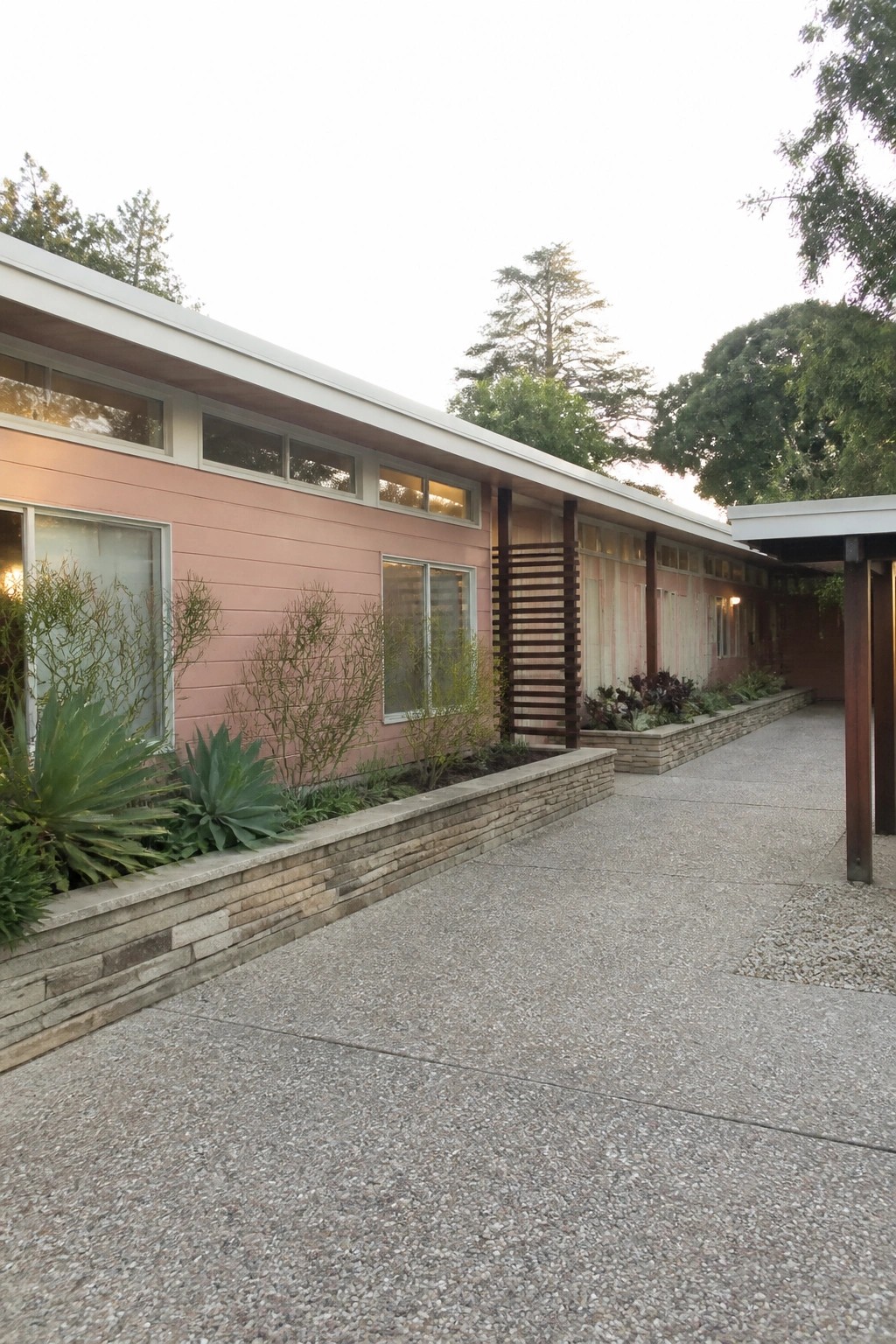 A single-story house with blush pink horizontal siding, white roof trim, and multiple windows set above a low stone retaining wall.
