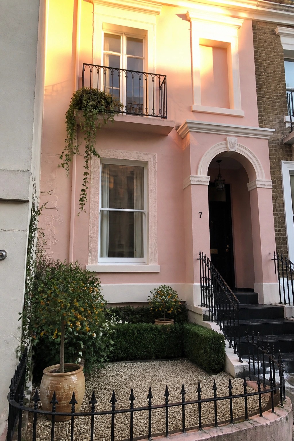 A two-story blush pink house exterior with warm white trim around windows and an arched doorway, black metal balcony, and black railings on the front steps.