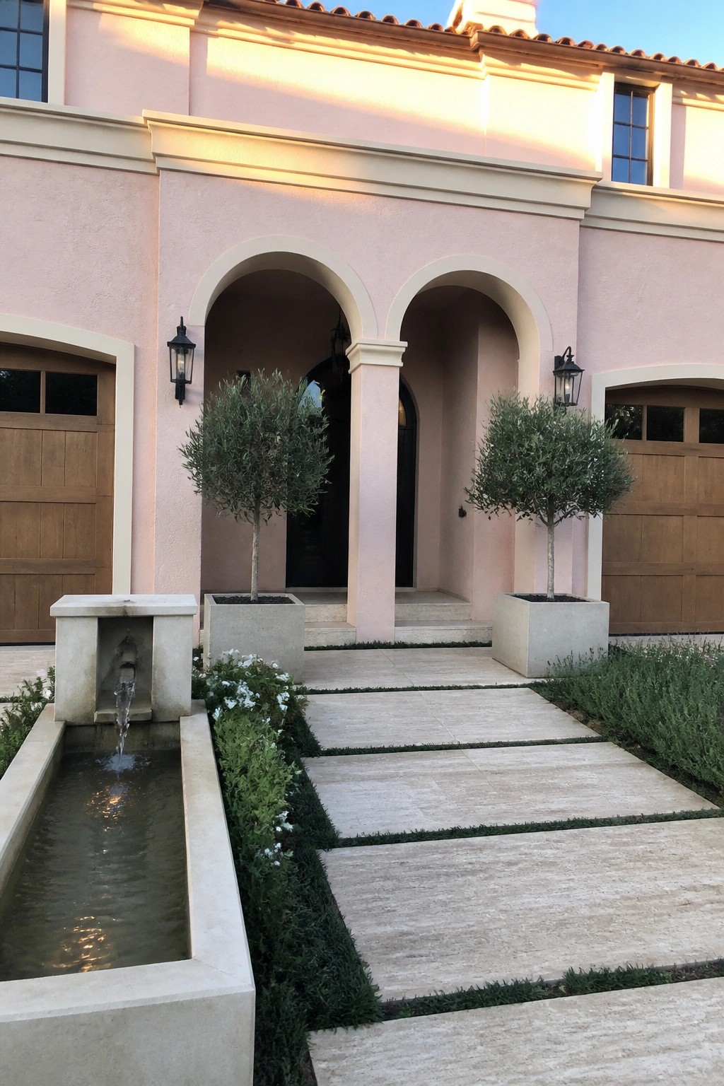 Blush pink stucco house exterior with warm white trim around arched openings, two potted trees, a small water feature on the left, and a stone pathway leading to the front door.