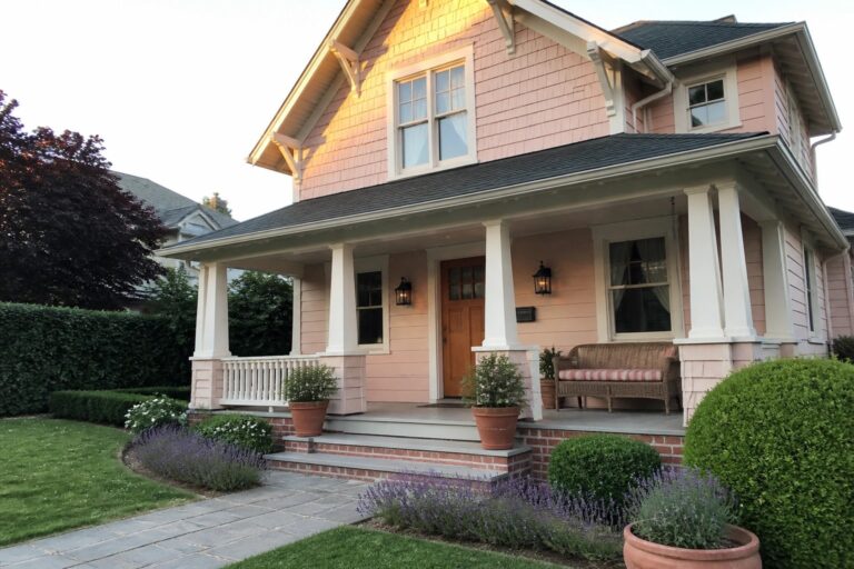A two-story pink house with white trim, a front porch with columns, wooden door, and stone pathway in a landscaped garden.