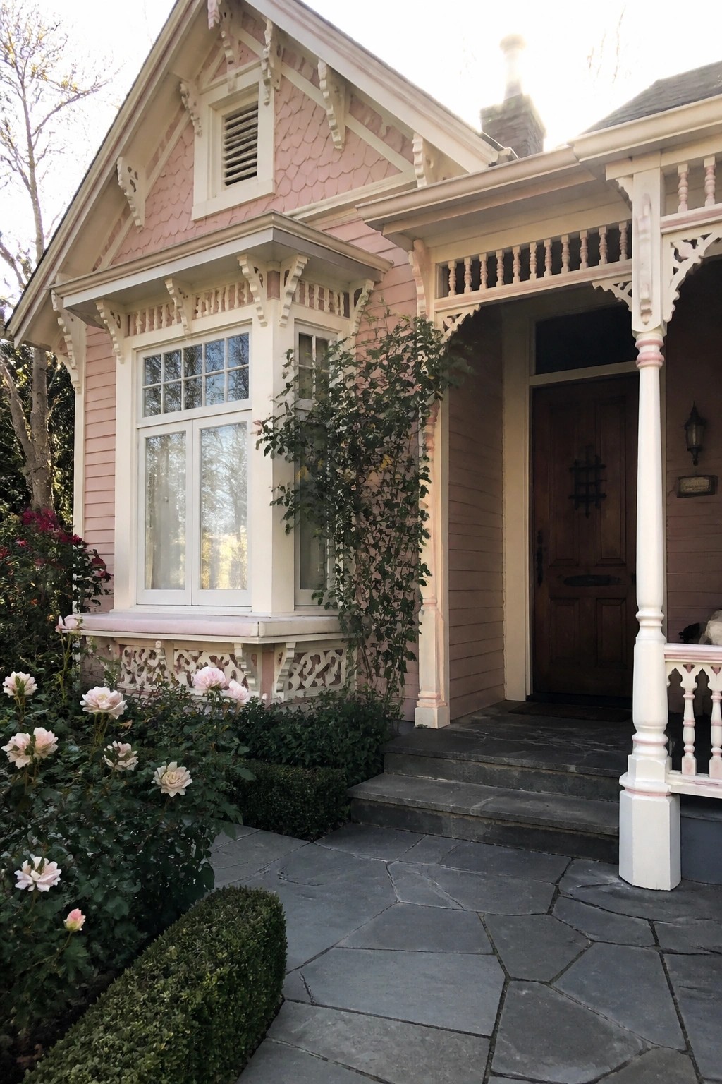 A blush pink house exterior with warm white trim, a covered porch, wooden front door, and rose bushes along the stone path.