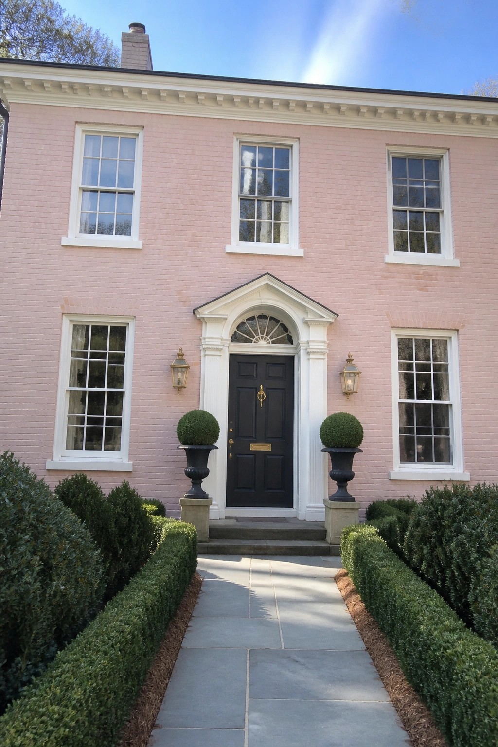 Pink brick house with black door, potted topiaries, and hedged stone walkway.