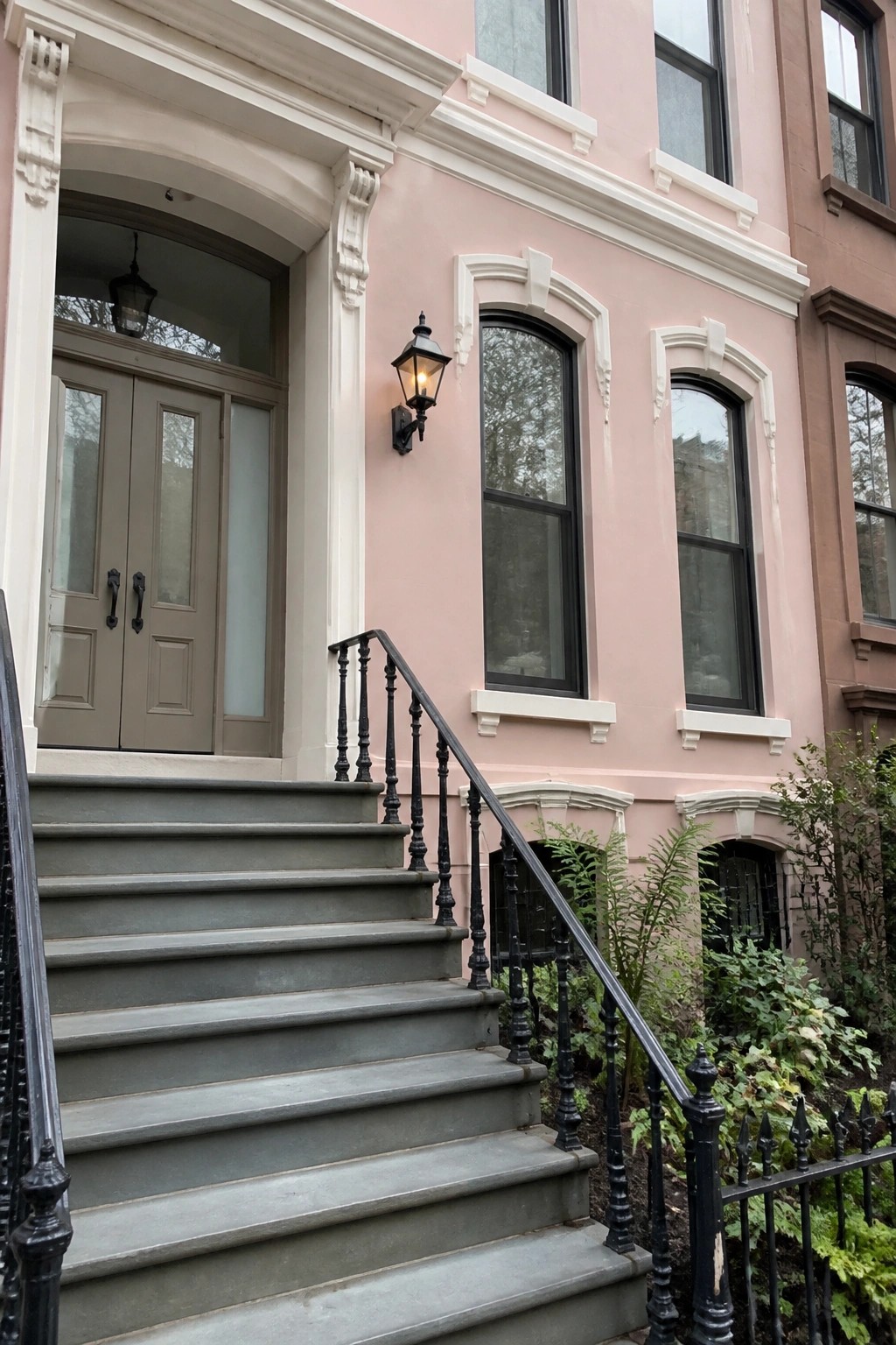 Pink townhouse entrance with gray stairs, black railings, arched windows, and lit lantern.