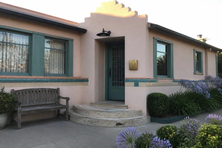 19 Playful Pink and Teal House Exteriors Inspired by Art Deco Color A light pink stucco house exterior with teal window frames, teal tile bands, a teal door, and a geometric stepped parapet above the entrance.