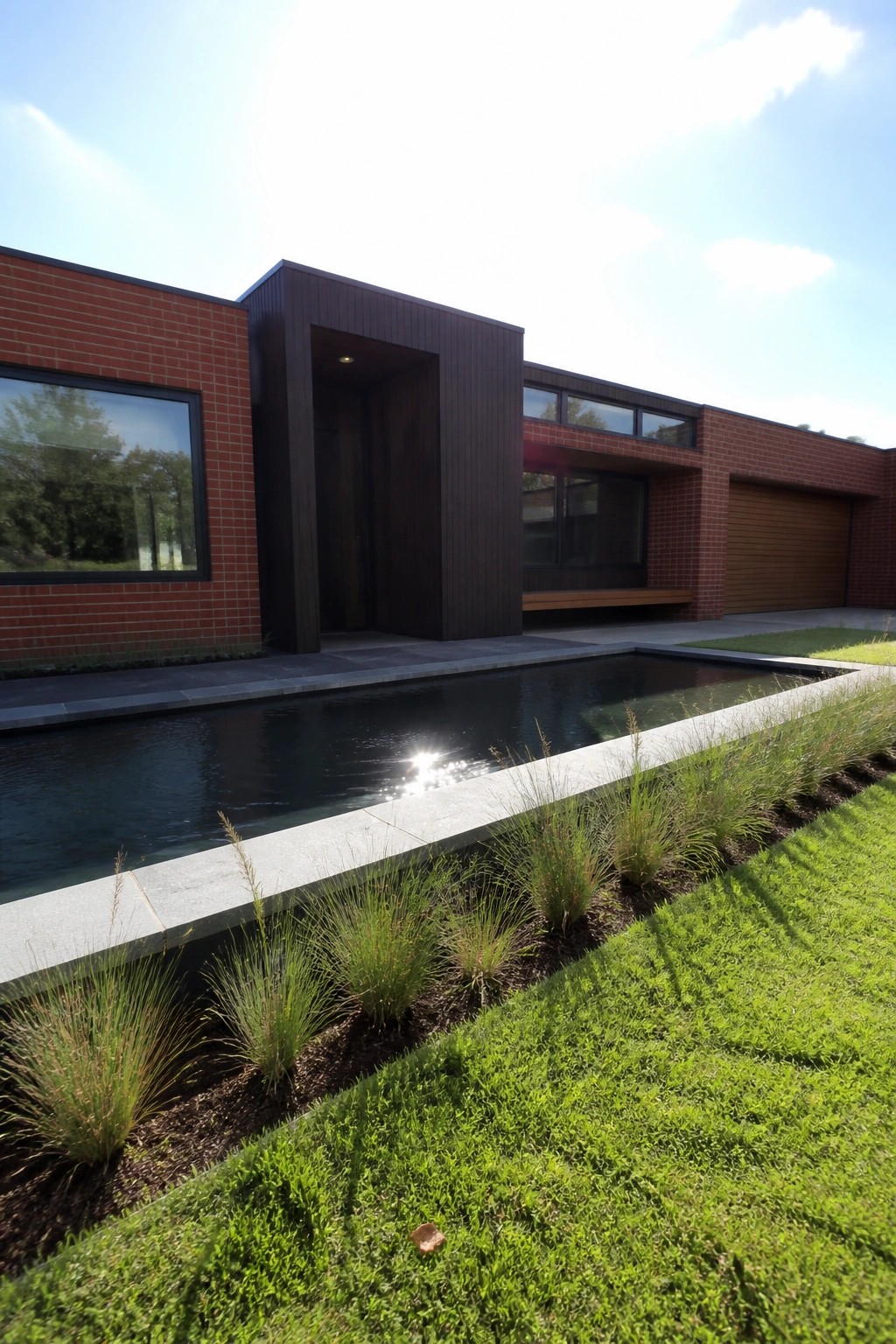 Modern red brick ranch exterior with a projecting dark wood entry volume, horizontal windows, and a long pool in the foreground.